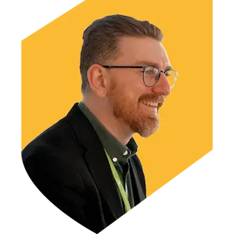 A man wearing glasses with a grey shirt and black jacket on a yellow shield background.