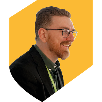 A man wearing glasses with a grey shirt and black jacket on a yellow shield background. 